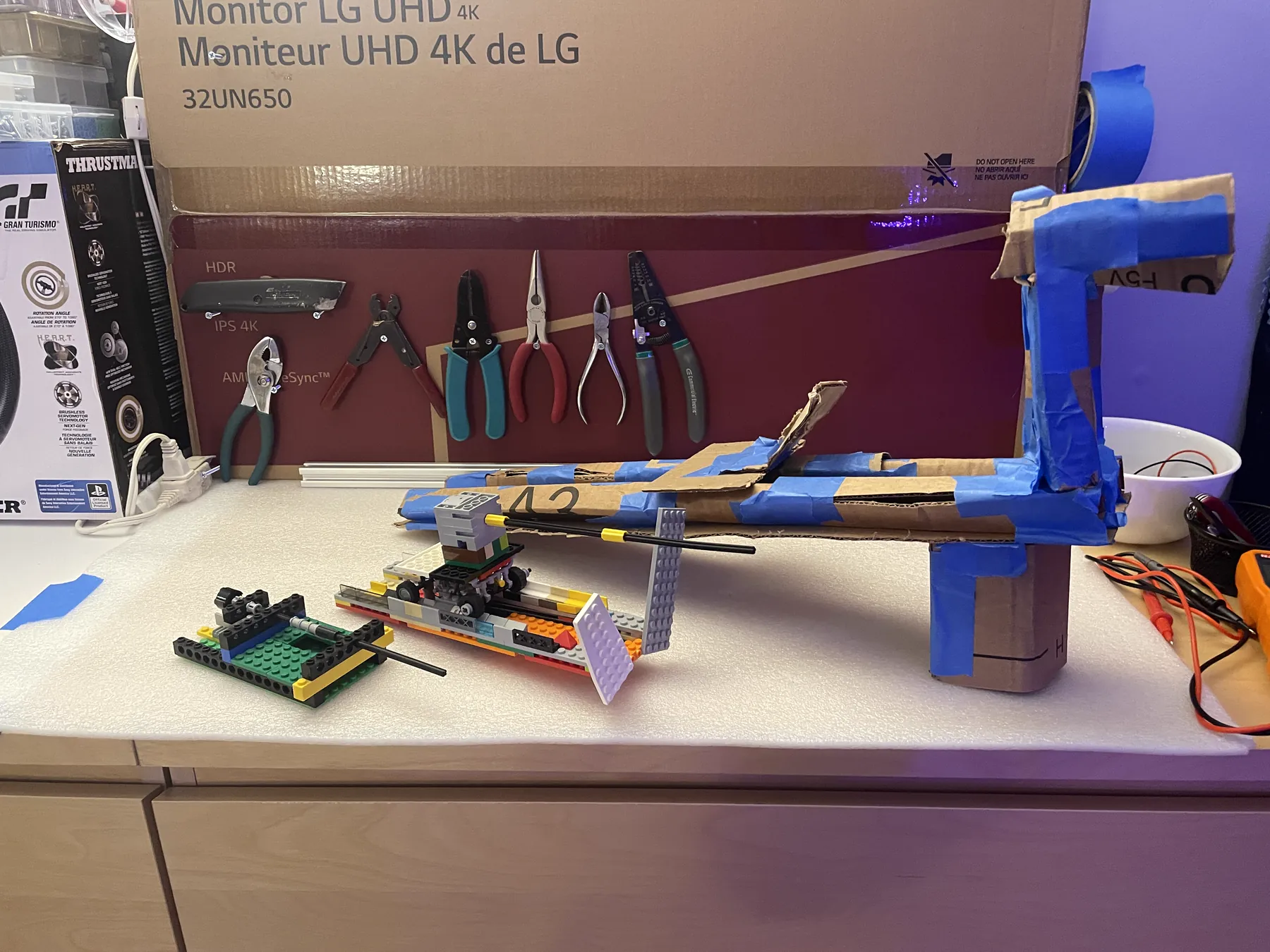 Prototype models of the goalie mechanism built from Legos and taped cardboard, on a counter in front of a pegboard of pliers and cutters