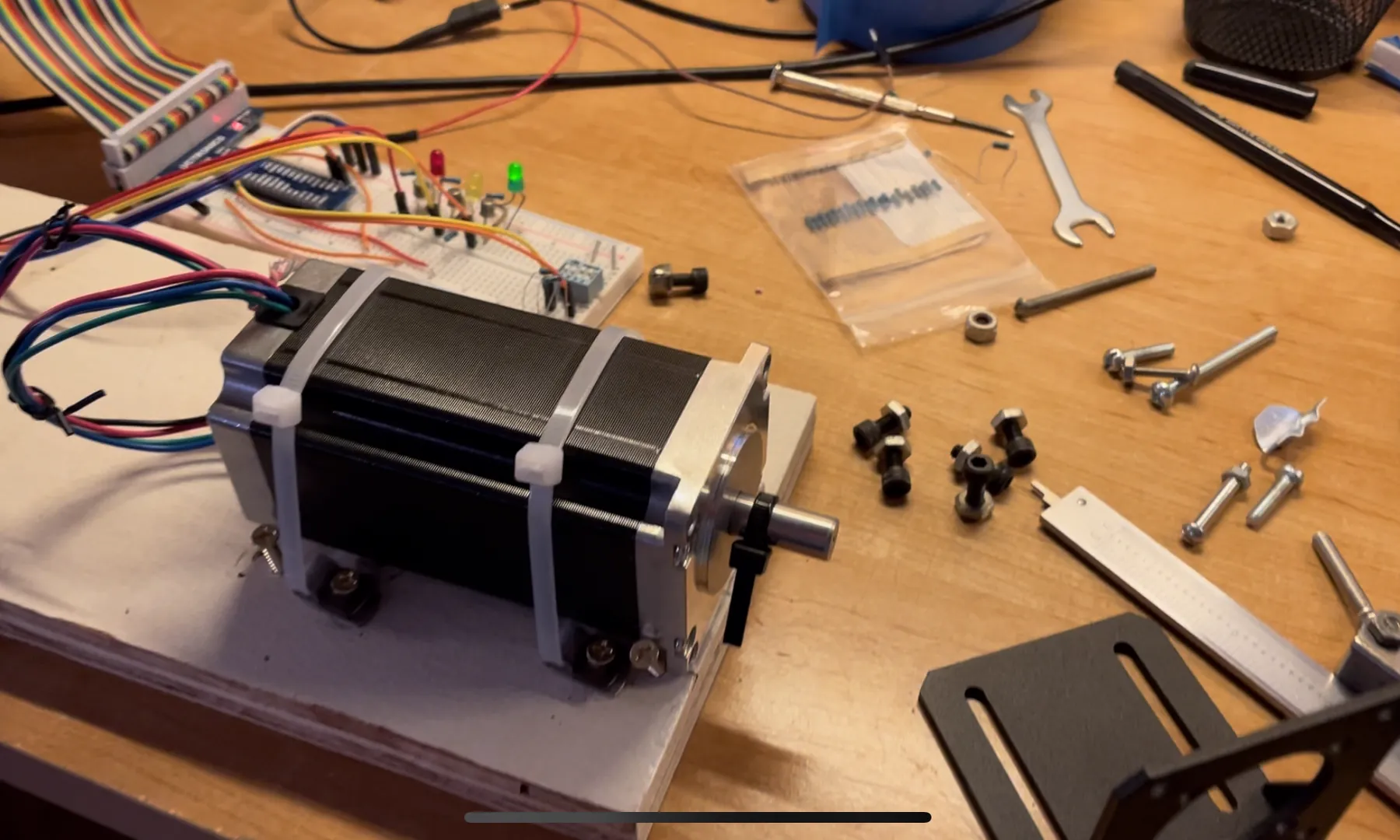 The stepper motor bolted to a board next to a breadboard with LEDs and a ribbon cable, surrounded by tools, bolts and mounting hardware